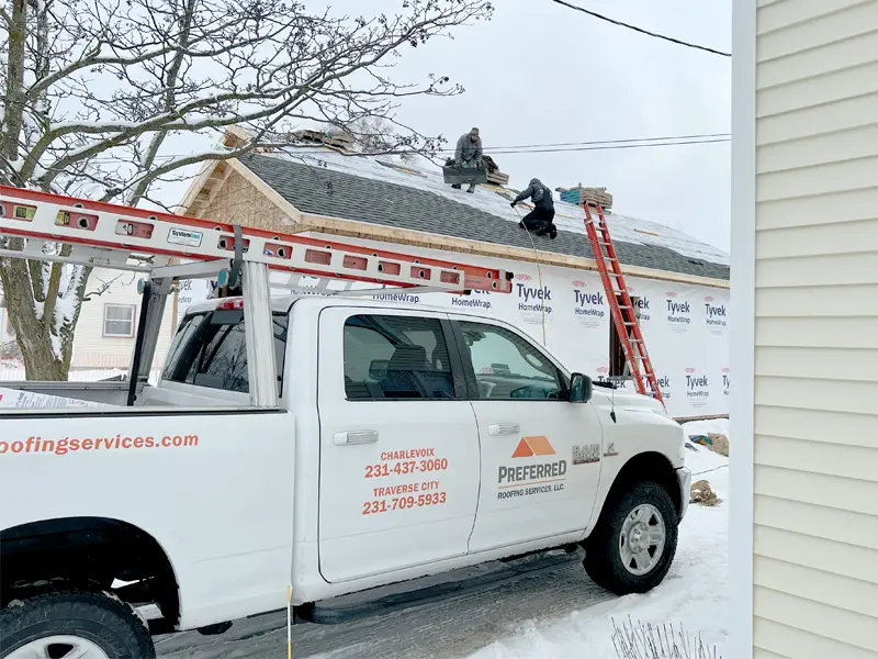 Photo of Preferred Roofing Services truck in front of a home with roofers on the roof replacing the shingles.