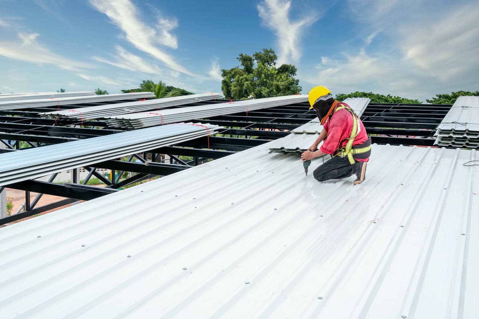 Worker in safety gear installing white metal roofing sheets on a building under a blue sky.