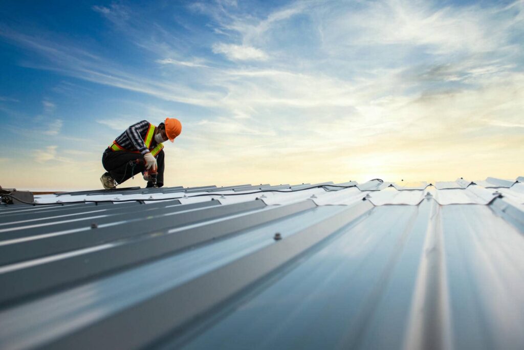 Construction worker wearing a hard hat and mask using a drill on a metal roof under a cloudy sky.