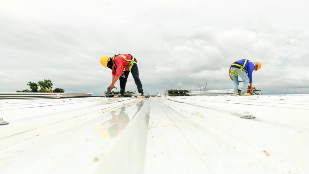 Two construction workers wearing helmets and harnesses working on a metal roof under a cloudy sky.