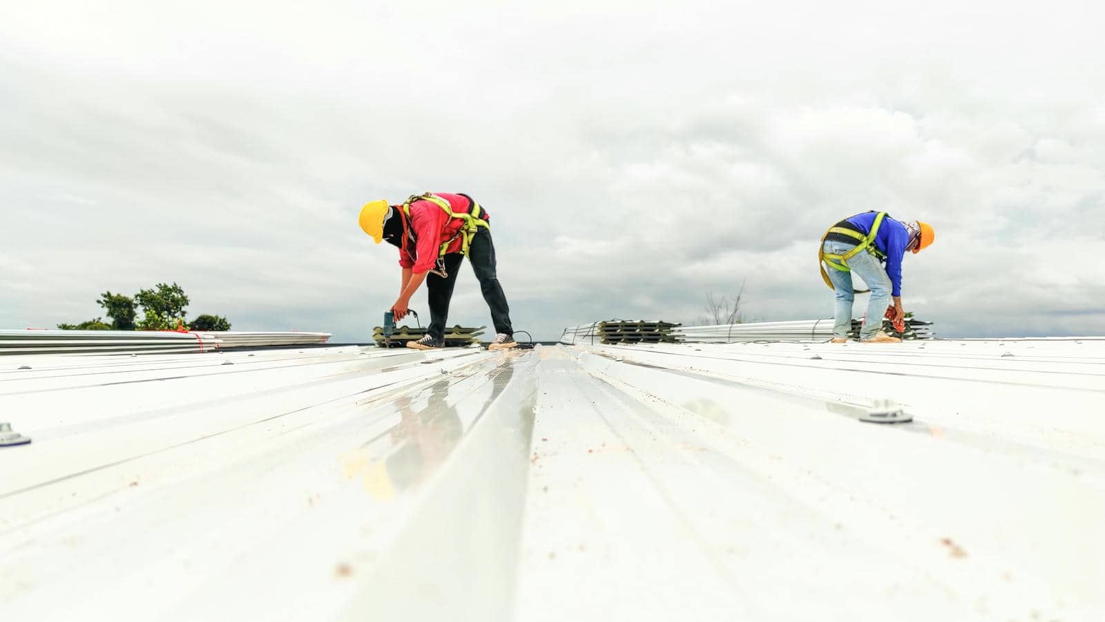 Two construction workers wearing helmets and harnesses working on a metal roof under a cloudy sky.