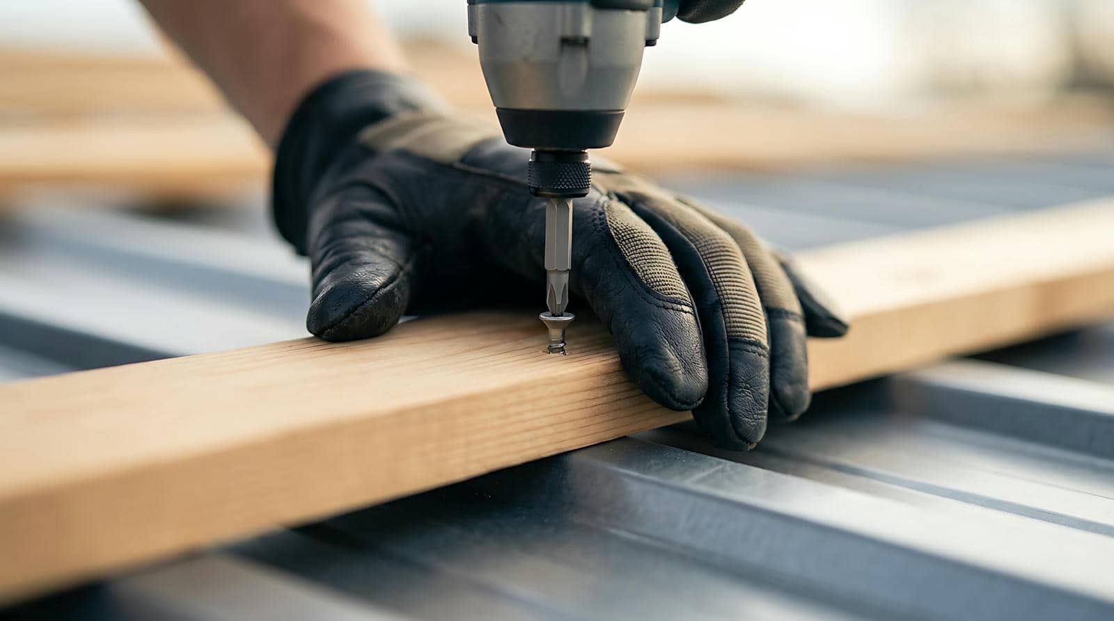 Hand in black glove using a power drill to screw into a wooden plank.