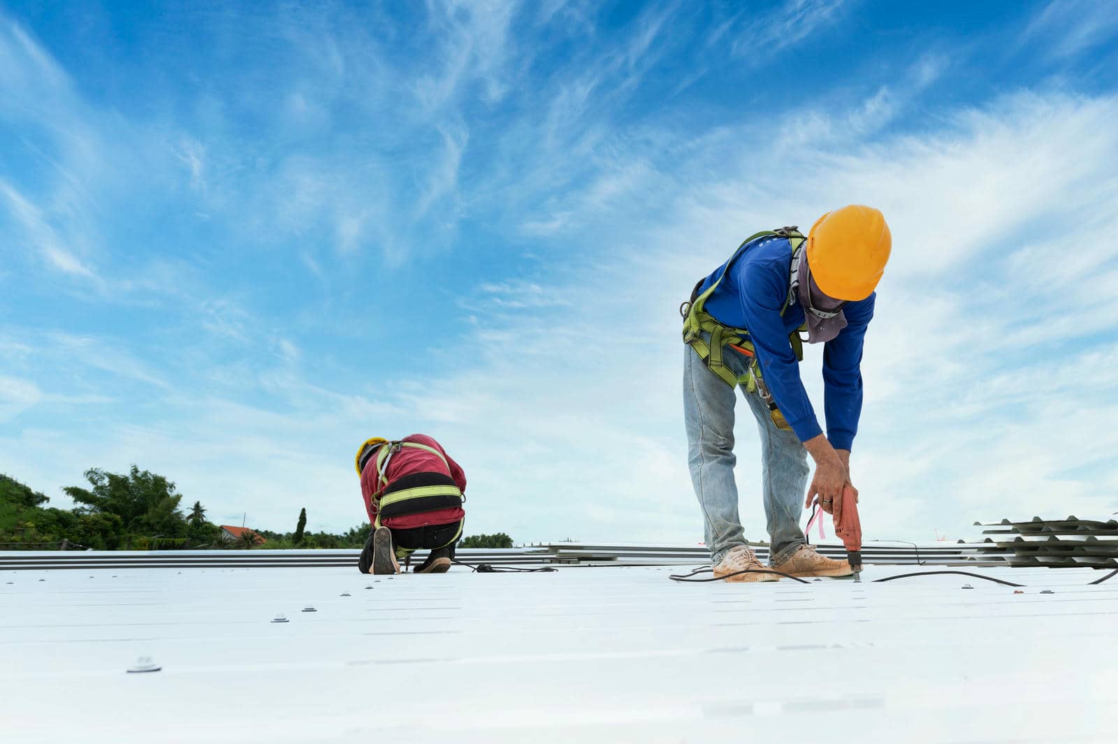 Two construction workers wearing safety harnesses and helmets working on a metal roof under a blue sky.