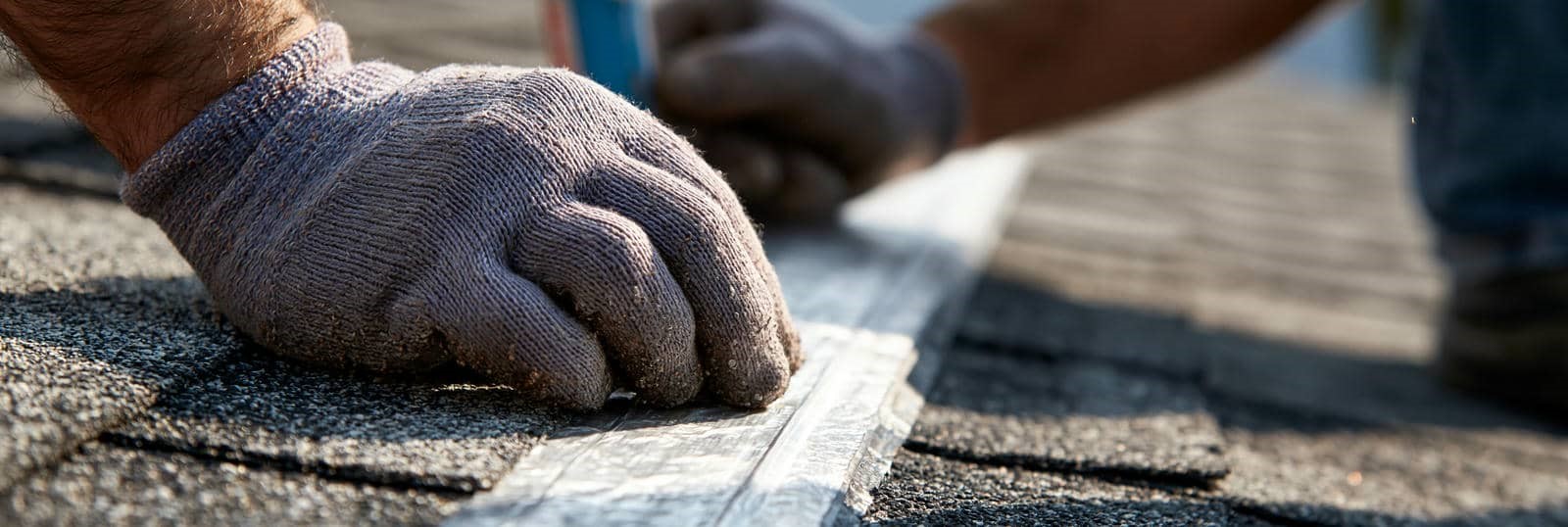 Gloved hands applying adhesive tape on asphalt roof shingles.