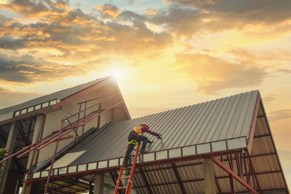 Worker in safety gear climbing a ladder to repair a metal roof at sunset.