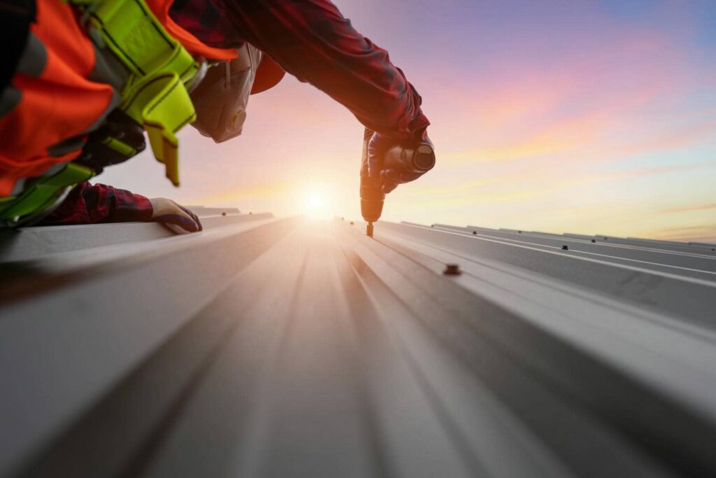 Worker wearing safety gear using a power drill on a metal roof at sunset.