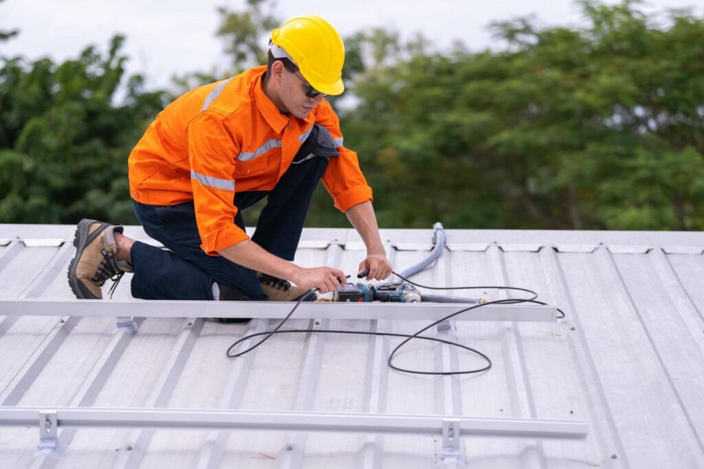 Worker in a yellow helmet and orange jacket kneels on a metal roof, handling electrical cables.
