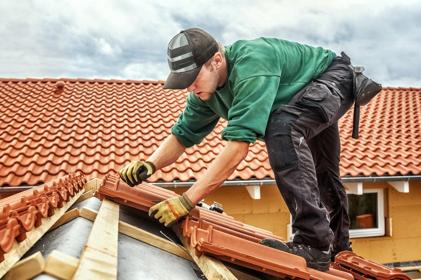 A man in a green sweatshirt and cap installs red roof tiles on a house.