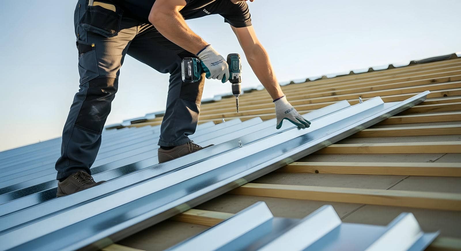 A worker installs metal roofing panels using a cordless drill on a wooden frame.