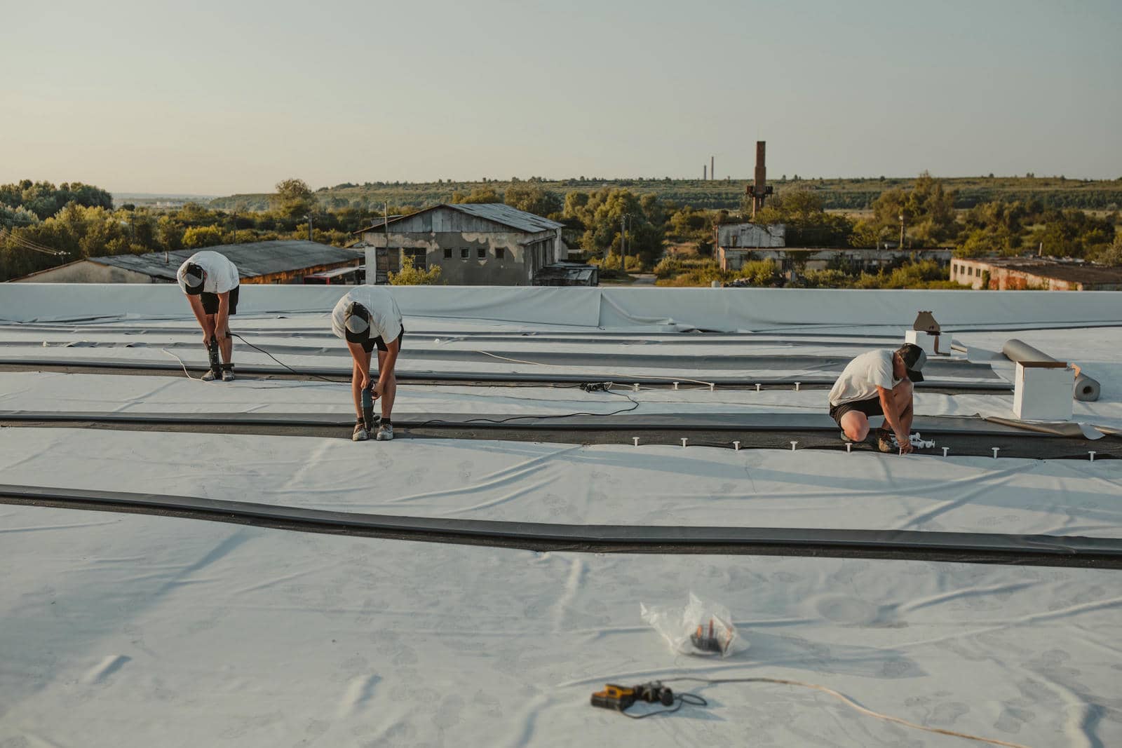 Three workers installing roofing material on a flat roof during daylight.