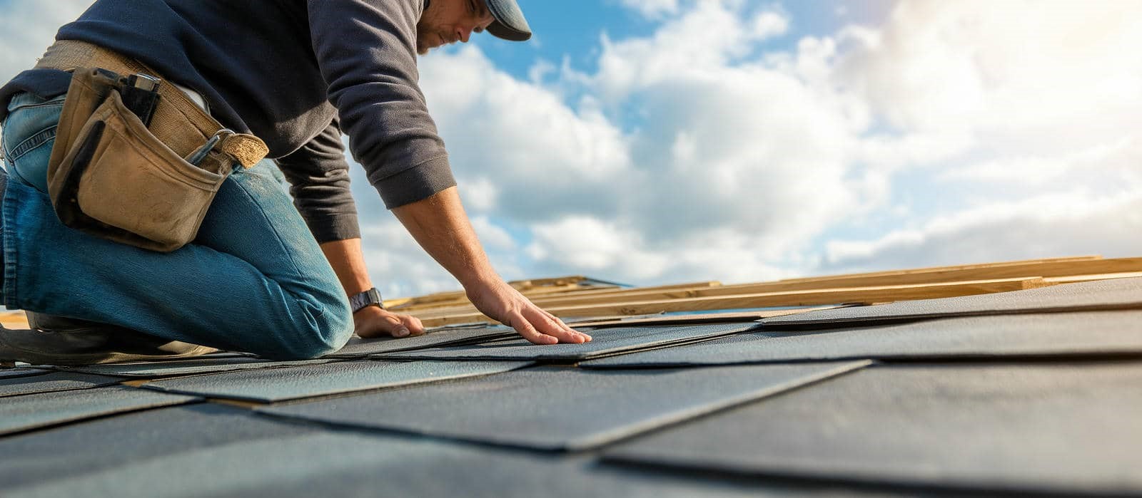 A person kneeling on a roof, inspecting or installing dark shingles under a partly cloudy sky.
