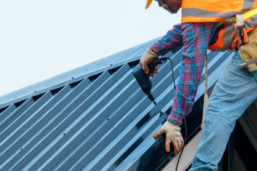 Construction worker in safety gear using a power drill on a metal roof.