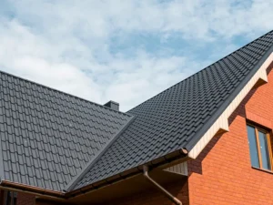 Close-up of a house roof with dark gray metal tiles and a brick wall under a partly cloudy sky.