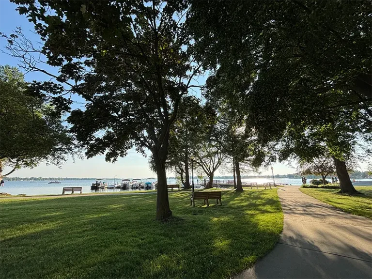Park pathway with benches and trees beside a calm lake under a clear sky.