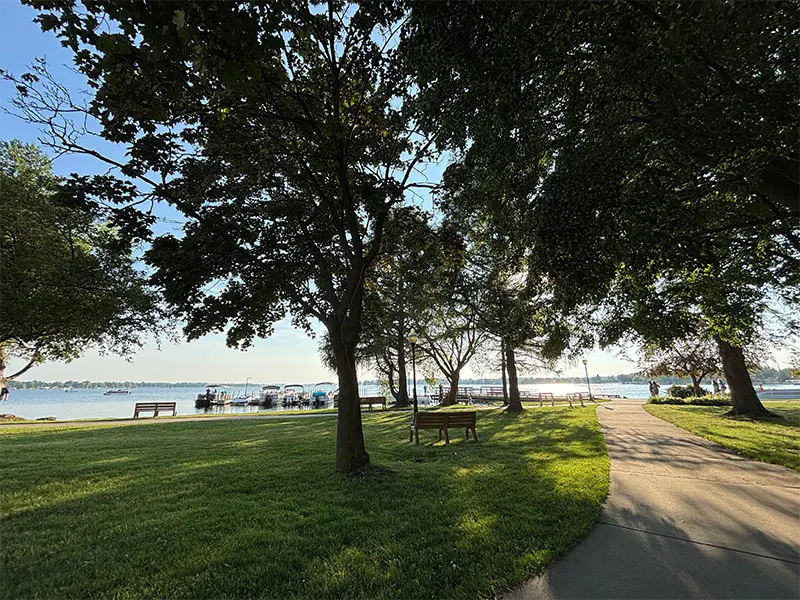 Park pathway with benches and trees beside a calm lake under a clear sky.