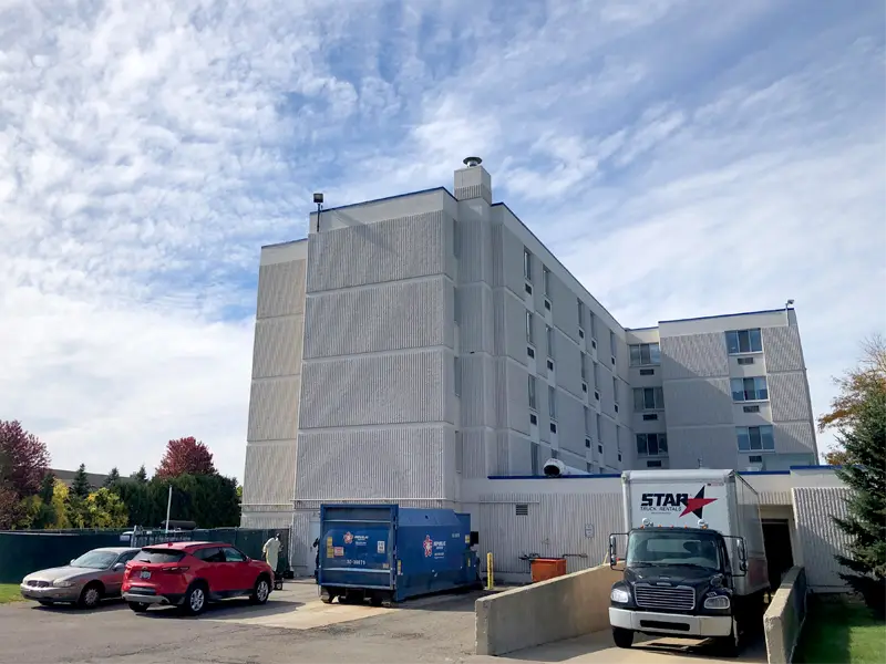 Large multi-story beige building with parked cars, a blue dumpster, and a white delivery truck in front.