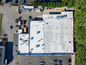 Aerial view of a large flat-roofed commercial building surrounded by parked cars and trees.