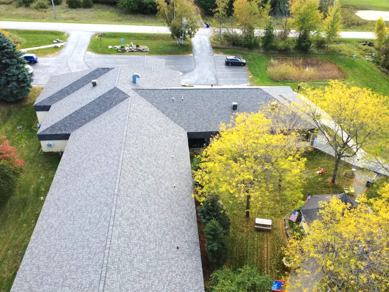 Aerial view of a large building with a gray roof surrounded by trees with yellow leaves and a small parking area.