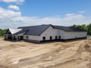 Large white and black metal building with a black roof on a dirt lot under a partly cloudy sky.