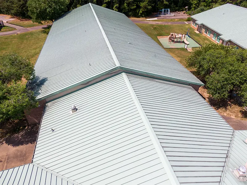 Aerial view of a large building with a light green metal roof and a playground nearby.