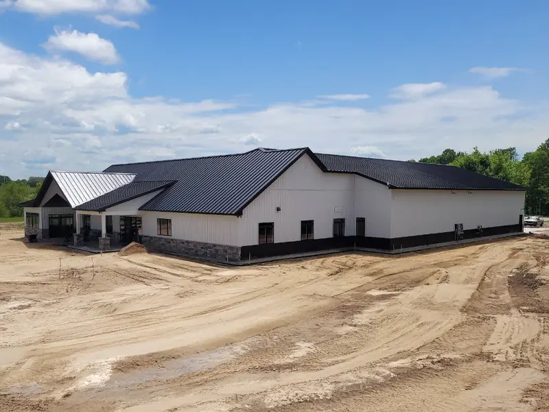 Large white and black metal building with a black roof on a dirt lot under a partly cloudy sky.
