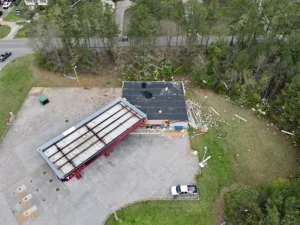 Abandoned gas station with damaged roof and debris scattered on the ground near a wooded area.