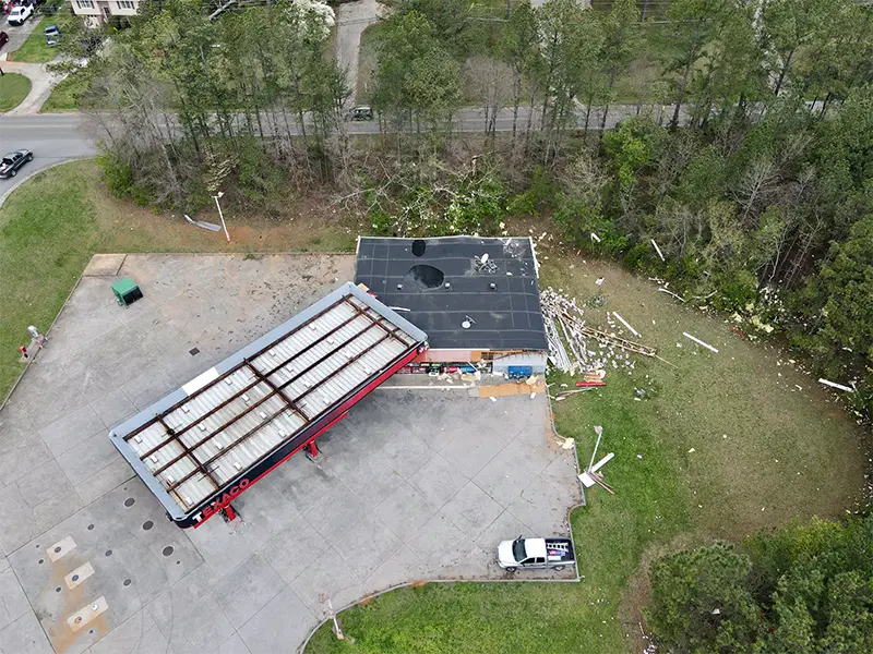 Abandoned gas station with damaged roof and debris scattered on the ground near a wooded area.