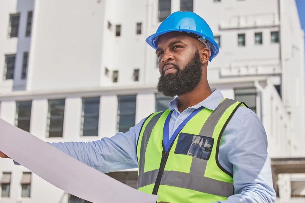 Construction worker in a blue hard hat and safety vest holding a blueprint at a building site.