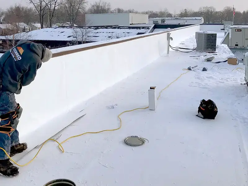 Worker in winter clothing inspecting or working on a flat snowy rooftop with HVAC units.