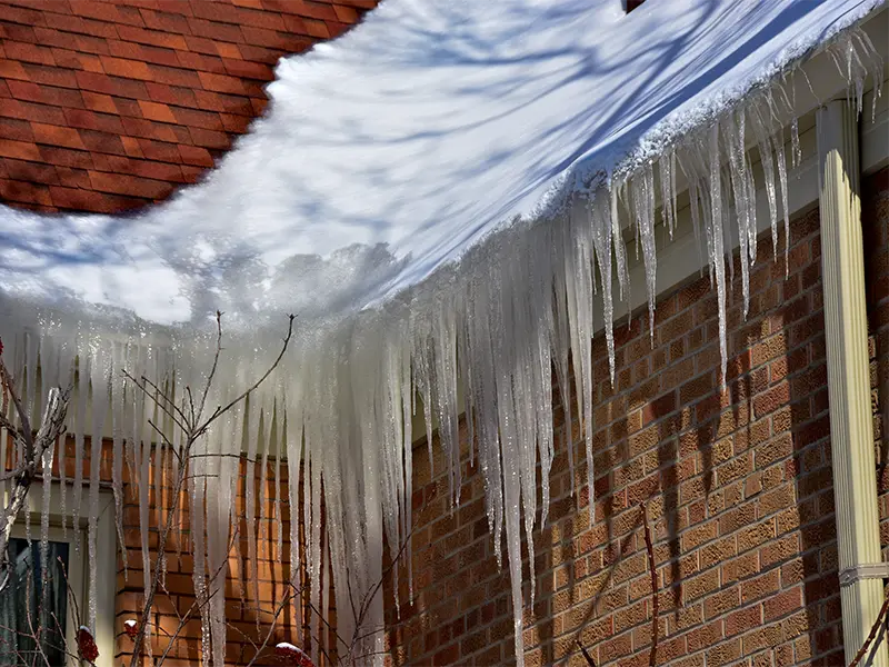 Long icicles hanging from a snow-covered roof edge above a brick wall.
