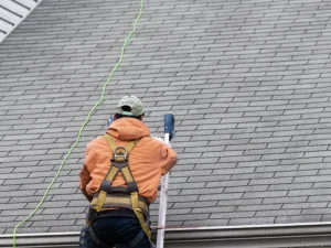 Worker in safety harness climbing a ladder against a shingled roof.