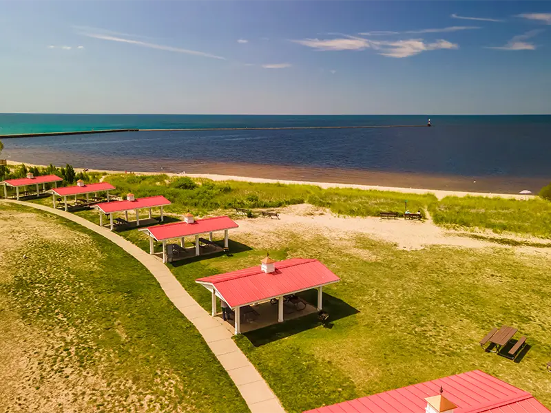 Red-roofed picnic shelters along a walkway near a sandy beach and calm ocean.
