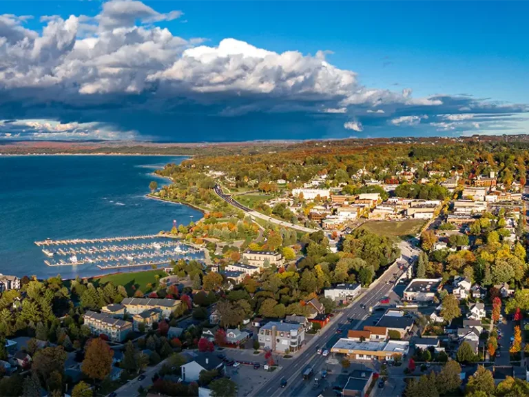 Aerial view of a lakeside town with a marina, roads, and autumn-colored trees under a partly cloudy sky.
