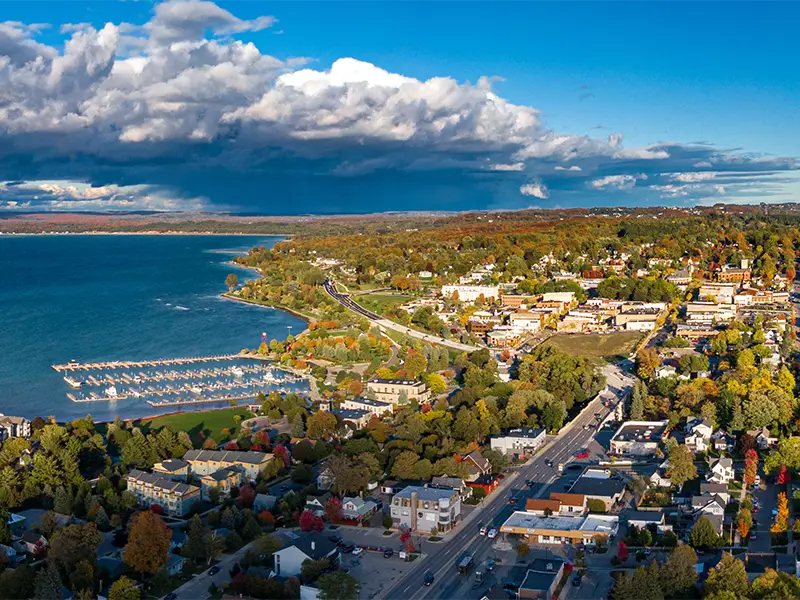 Aerial view of a lakeside town with a marina, roads, and autumn-colored trees under a partly cloudy sky.
