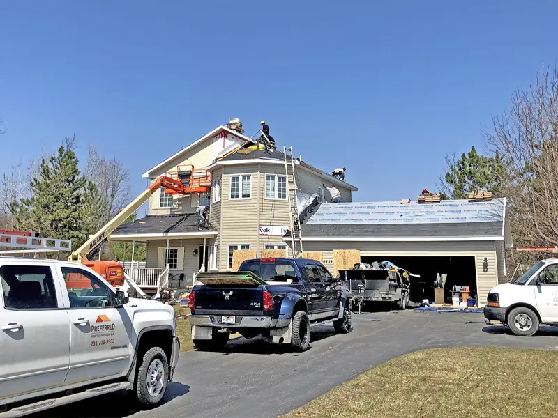 Workers on a house roof under construction with trucks and equipment in the driveway.