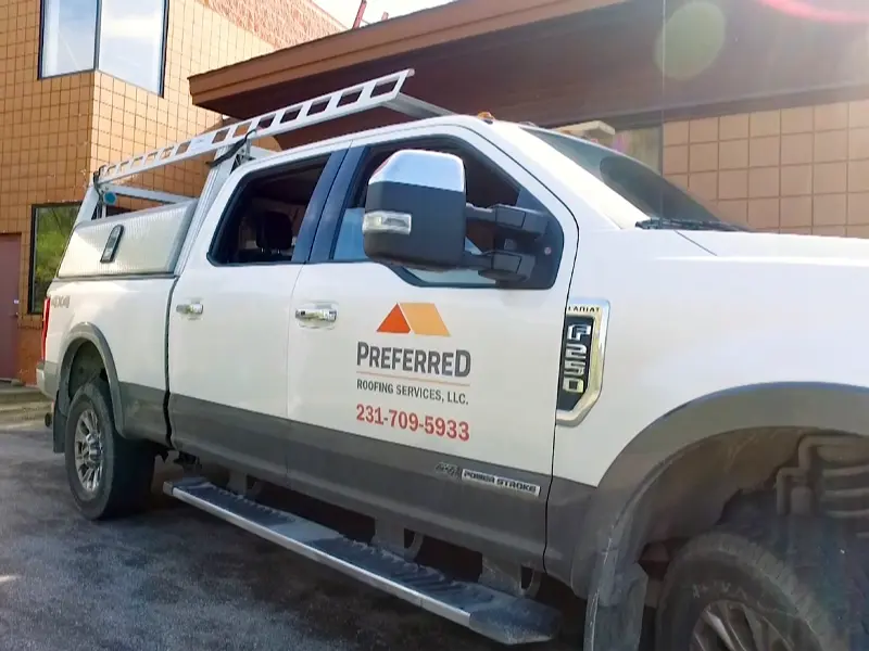 White pickup truck with ladder and "Preferred Roofing Services" logo parked outside a building.