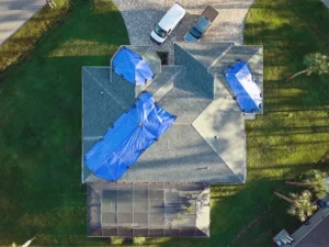 Aerial view of a house with blue tarps covering damaged roof sections and two vehicles parked in the driveway.
