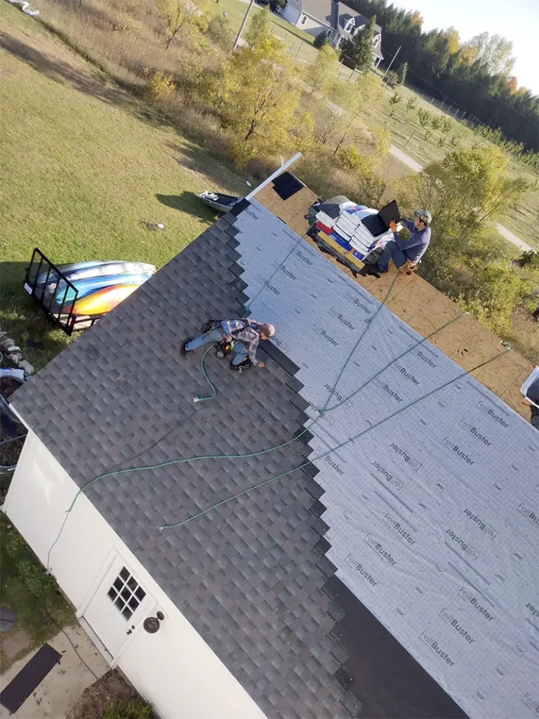 Two workers installing shingles on a roof, one sitting near roofing materials and the other nailing shingles.