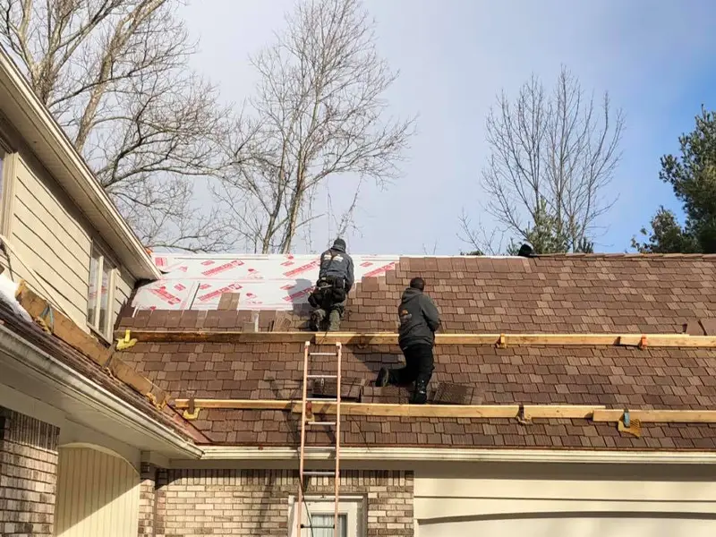 Two workers installing brown shingles on a roof with wooden scaffolding and a ladder.
