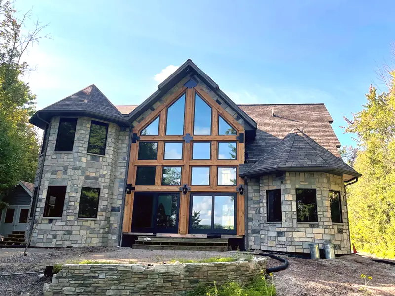 Stone house with large triangular wooden-framed windows and a shingled roof surrounded by trees.